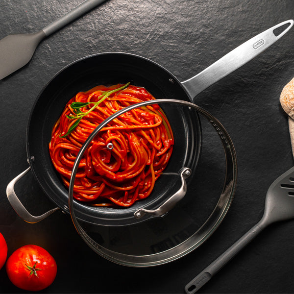 A pot of cooked spaghetti in rich tomato sauce, garnished with a sprig of rosemary, sits on a dark surface with a glass lid partially covering it. Two spatulas and fresh tomatoes are placed nearby.