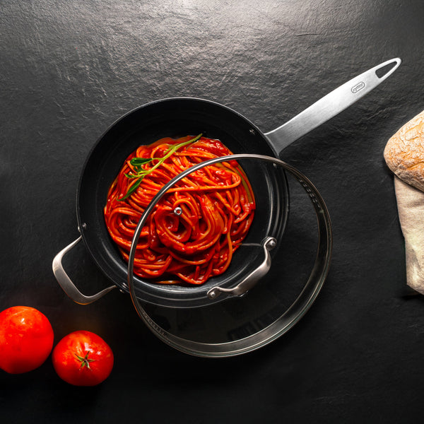 A pan of spaghetti with tomato sauce and basil sits on a black surface. A glass lid partially covers the Zyliss Ultimate Pro Ceramic Saute Pan With Lid, prized for its non-stick coating. Two tomatoes and ciabatta bread wrapped in cloth are nearby. 