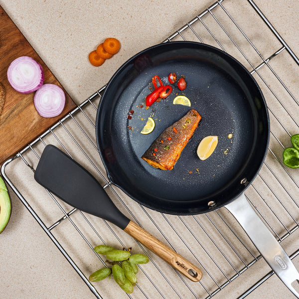 A cooked salmon fillet with lemon and red chili sits in a black pan on a cooling rack, surrounded by onion, carrot, avocado, lime, and grapes. The Zyliss Oak Wood Easy Lift Turner rests beside the pan.