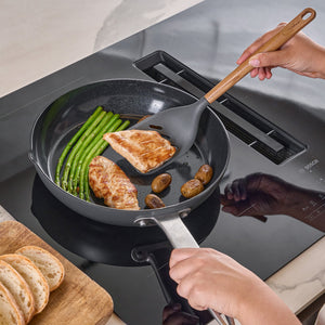 A person uses the Zyliss Oak Wood Slotted Turner to cook chicken breast, green beans, and small potatoes in a black frying pan on the stovetop, with slices of bread resting on an oak board for a classic kitchen moment.