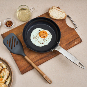 A fried egg with pepper flakes in a frying pan, beside the Zyliss Oak Wood Slotted Turner, bread, chili flakes, oil cup, and a plate of food on a beige countertop—a kitchen essential setup.