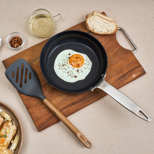 A fried egg with pepper flakes in a frying pan, beside the Zyliss Oak Wood Slotted Turner, bread, chili flakes, oil cup, and a plate of food on a beige countertop—a kitchen essential setup.