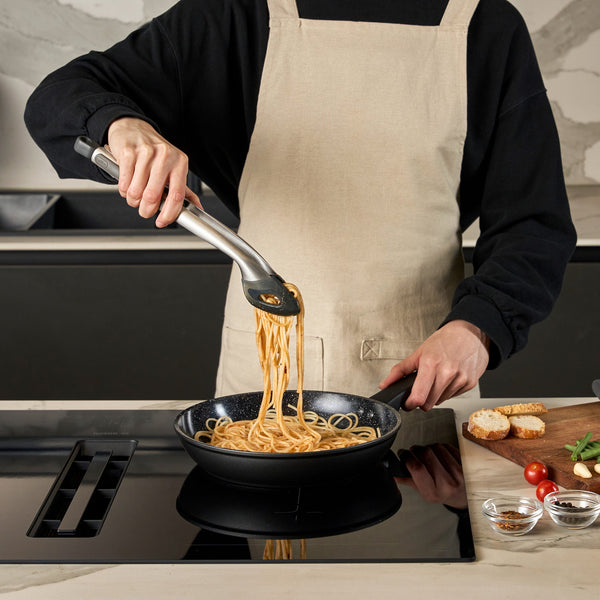 #20cm/8"
Wearing a beige apron and black shirt, a person uses tongs to serve pasta from the Zyliss Ultimate Ceramic Fry Pan on a stovetop, with small bowls of ingredients and slices of bread displayed nearby.