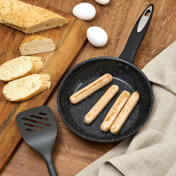 #20cm/8"
The Zyliss Ultimate Ceramic Fry Pan with four sausages is on a wooden table alongside a spatula, sliced bread, whole eggs, and a loaf of bread on a board, with a beige cloth nearby.