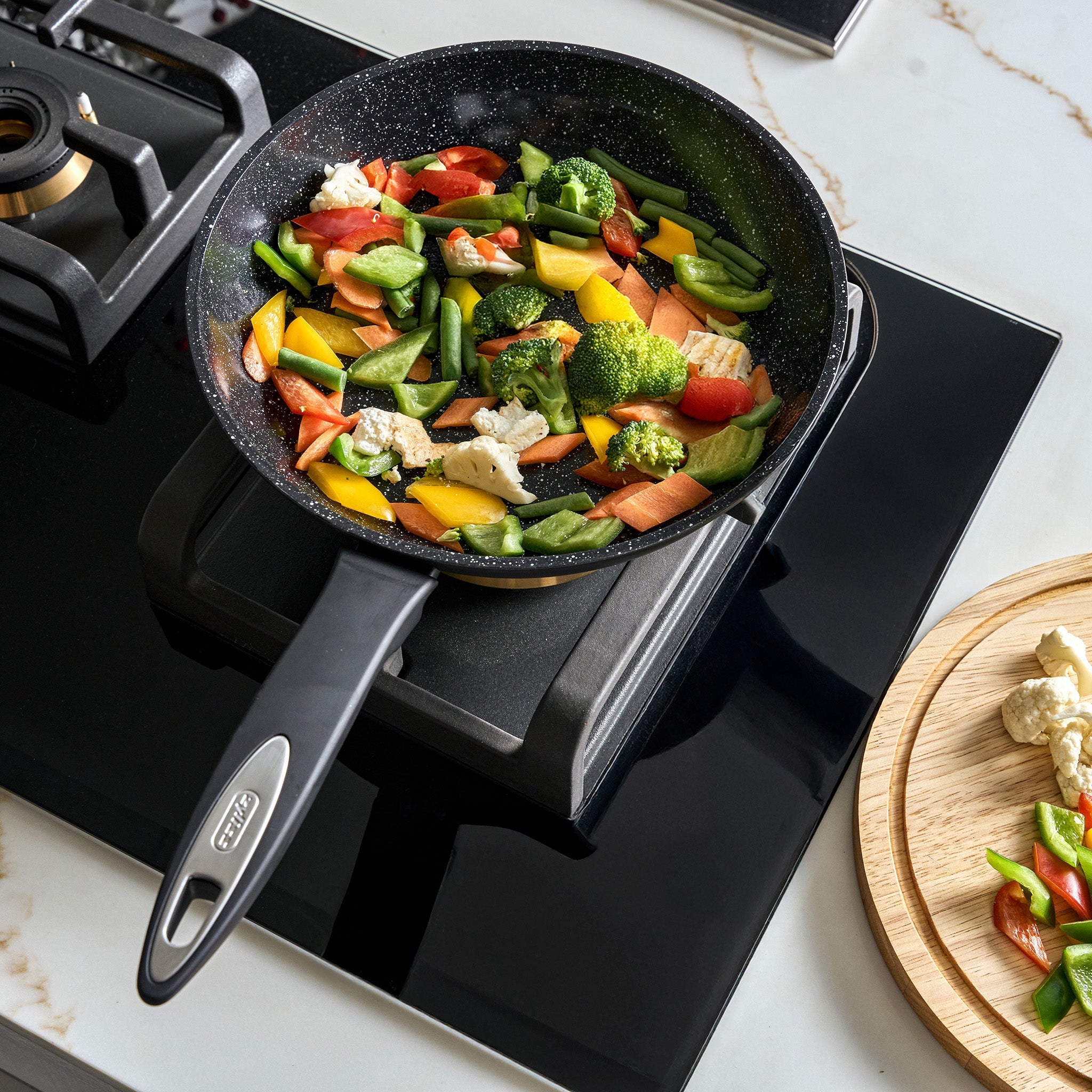 #24cm/9½"
The Zyliss Ultimate Ceramic Fry Pan cooks a vibrant mix of broccoli, carrots, and bell peppers on an induction stovetop, while extra veggies wait on a wooden cutting board—perfect for preparing healthy meals.