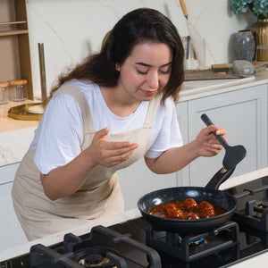 #24cm/9½"
A woman in a white shirt and beige apron leans over a stove, smelling food in her Zyliss Ultimate Ceramic Fry Pan, while holding a spatula in her modern kitchen.