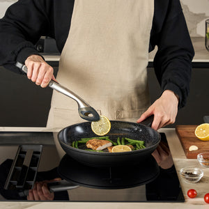 #28cm/11"
Wearing a beige apron, someone cooks fish and green beans in the Zyliss Ultimate Ceramic Fry Pan—an induction-compatible non-stick pan—for easy cooking and cleanup, topping with lemon using tongs. Nearby, a cutting board holds lemon, garlic, and spices.