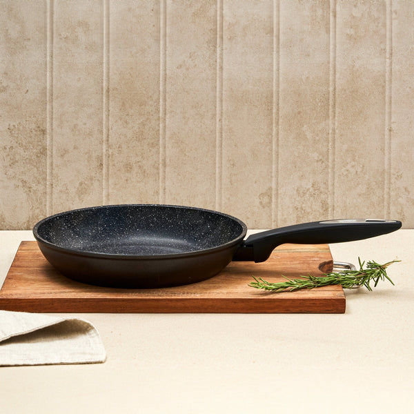 #28cm/11"
The Zyliss Ultimate Ceramic Fry Pan, featuring a black non-stick finish with a speckled interior, rests on a wooden cutting board beside fresh rosemary sprigs and a beige textured backsplash.