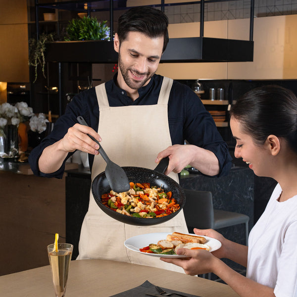 #28cm/11"
A smiling man in an apron serves a woman vibrant stir-fry from the Zyliss Ultimate Ceramic Fry Pan onto her plate as she sits at a table with a drink and salad in a modern kitchen.