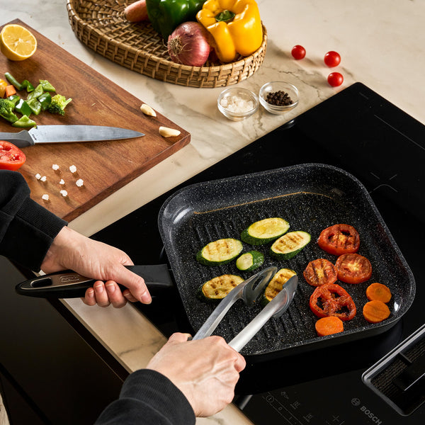 Using tongs, a person grills zucchini, tomato, and carrot slices in the Zyliss Ultimate Ceramic grill pan on a stovetop, with a cutting board of chopped veggies and a basket of whole vegetables nearby.