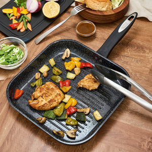 The Zyliss Ultimate Ceramic grill pan with a non-stick coating sits on a wooden table, filled with grilled chicken, assorted veggies, and mushrooms. Metal tongs rest on the induction-suitable pan beside bowls of greens and a sandwich.