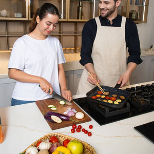 A woman slices veggies as a man in an apron cooks them on the stove using the Zyliss Ultimate Ceramic grill pan. A basket of fruits and vegetables rests on the kitchen counter.