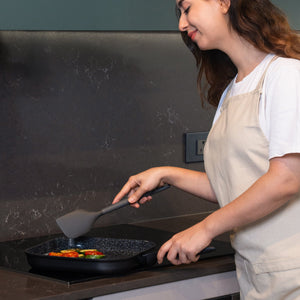 A woman in a beige apron smiles while cooking vegetables with a spatula in her modern kitchen, using the Zyliss Ultimate Ceramic grill pan—featuring a non-stick coating and induction stovetop compatibility.