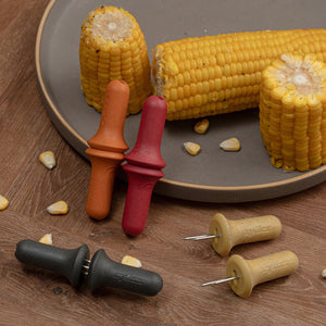 Three cooked corn on the cob pieces rest on a gray plate, each secured with a Zyliss Corn Holders 4 Piece Set featuring ergonomic handles and stainless steel pins, displayed on a wooden surface with scattered kernels.