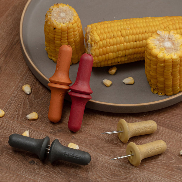 Three cooked corn on the cob pieces rest on a gray plate, each secured with a Zyliss Corn Holders 4 Piece Set featuring ergonomic handles and stainless steel pins, displayed on a wooden surface with scattered kernels.