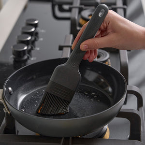 A hand uses the Zyliss Silicone Pastry Brush to spread oil in a nonstick frying pan on a four-burner gas stove.