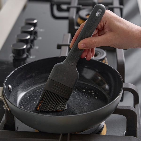 A hand uses the Zyliss Silicone Pastry Brush to spread oil in a nonstick frying pan on a four-burner gas stove.