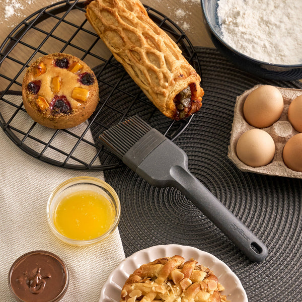 A black mat displays baked goods—a fruit tart, a pastry roll, and an almond pastry—surrounded by eggs, flour, melted butter, and chocolate spread. A Zyliss Silicone Pastry Brush is placed at the center.