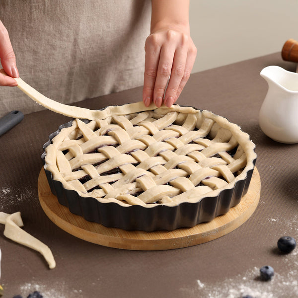 Someone weaves pie dough strips into a lattice over dark berries in the Zyliss Non-Stick Tart Pan with removable base, which sits on a wooden board on a brown table.