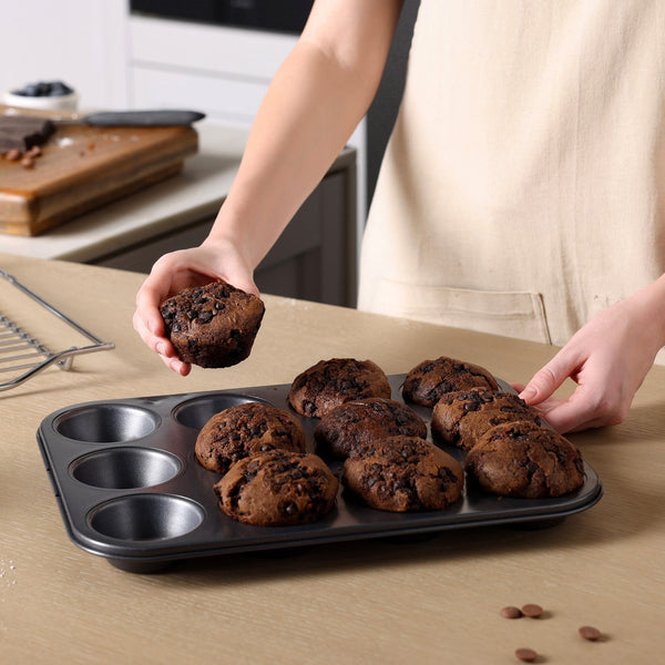 A person in a beige apron holds a chocolate muffin over the Zyliss Non-Stick 12 Hole Muffin Pan on the kitchen counter, with more muffins inside. A cooling rack and a wooden board with chocolate chips are nearby.