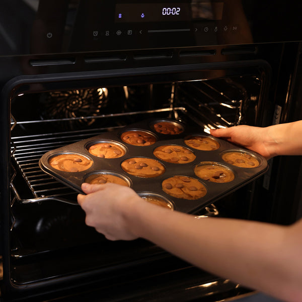 A person places the Zyliss Non-Stick 12 Hole Muffin Pan filled with chocolate chip batter into the oven to bake treats.