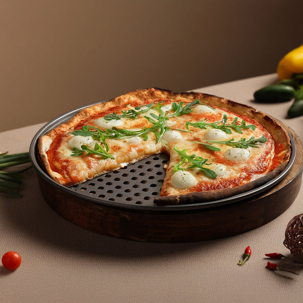 A round pizza topped with cheese, tomato sauce, and arugula rests on the Zyliss Non-Stick Pizza Tray with crispy bases. One slice is missing. Fresh tomato and chili are placed beside the tray.