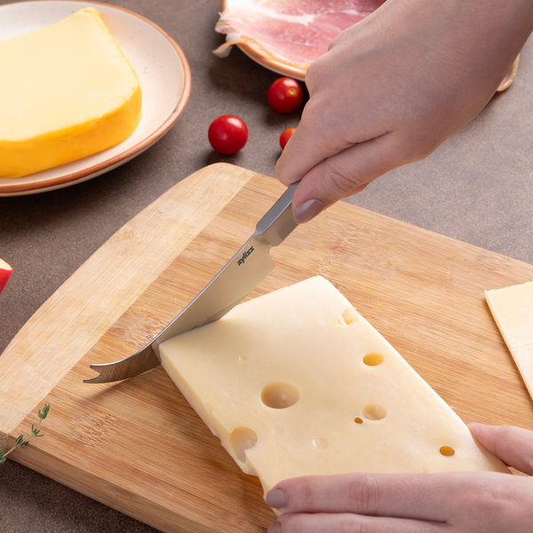 Using the Zyliss Signature Universal Cheese Knife, a person slices Swiss cheese on a wooden cutting board. Nearby are a wedge of yellow cheese, slices of meat, and cherry tomatoes.