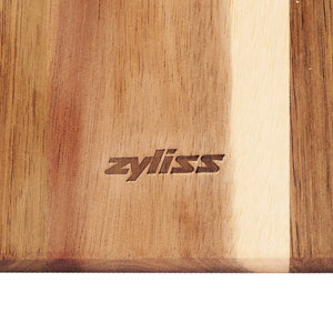 A close-up of the Signature 3 Piece Cheese Knife & Acacia Serving Board Set by Zyliss, featuring zyliss engraved in the center and showcasing striking light and dark brown acacia wood tones.