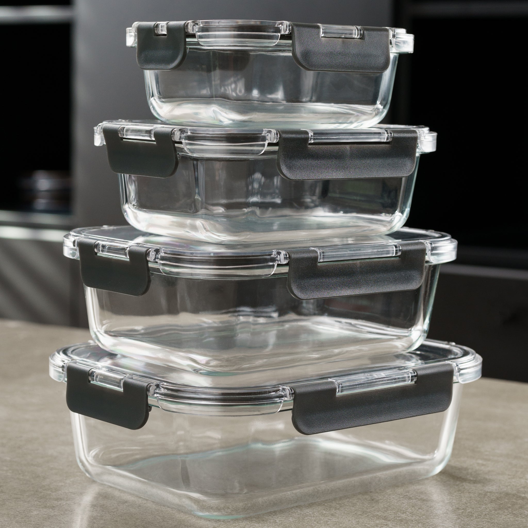 Four rectangular glass food storage containers with black locking lids are stacked on a kitchen countertop. The containers are clear, and the background is blurred, suggesting a modern kitchen setting.