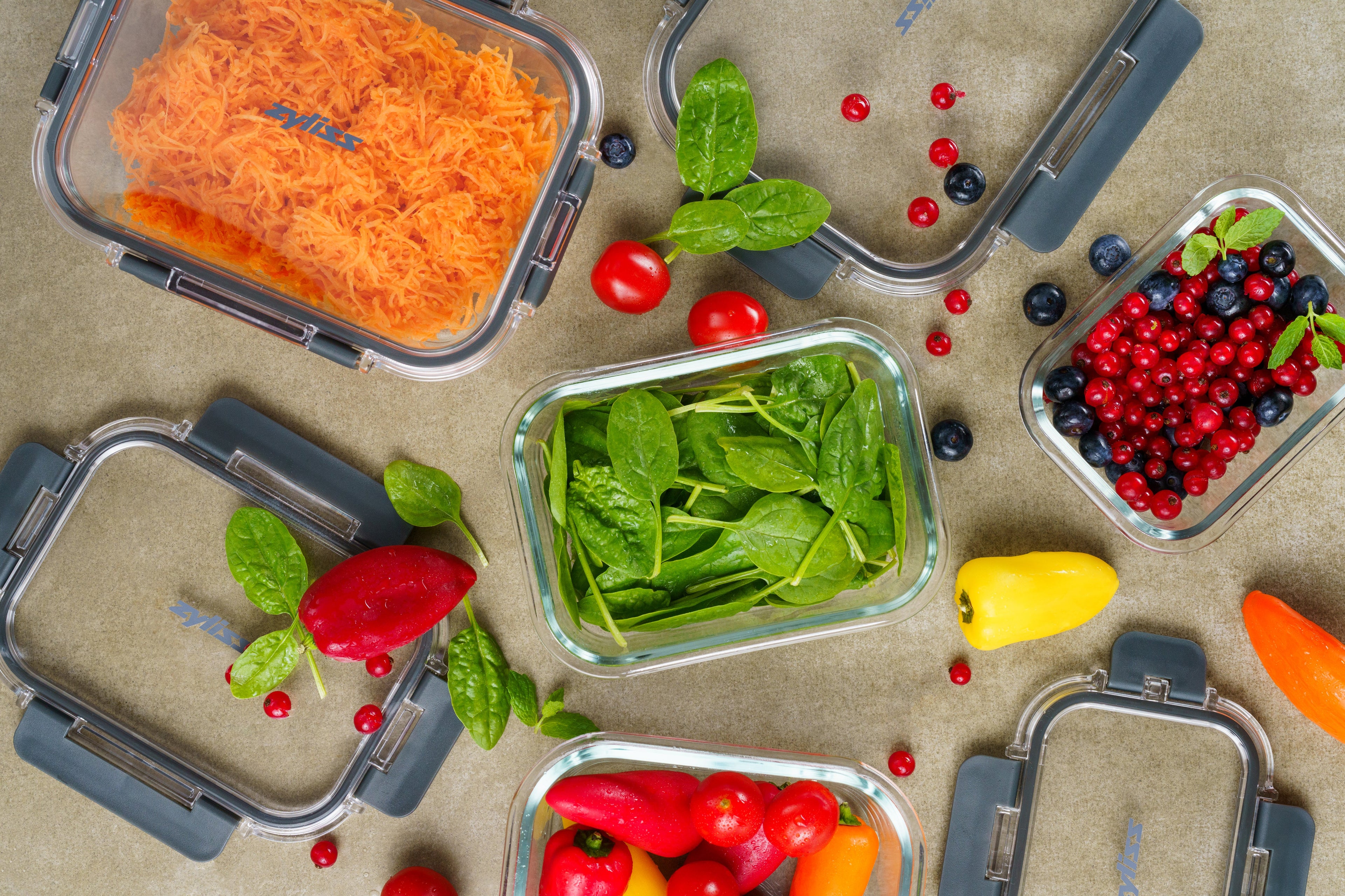 Glass food containers filled with shredded carrots, spinach, cherry tomatoes, colorful mini peppers, blueberries, and red currants are arranged on a countertop, some with lids off and some partially open.