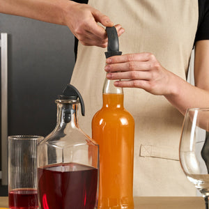 A person in a beige apron opens an orange bottle with a bottle opener beside a glass pitcher of red liquid, a glass, and an empty wine glass on wood—ideal for keeping drinks fresh with the Zyliss Bottle Stopper 2 Piece Set.