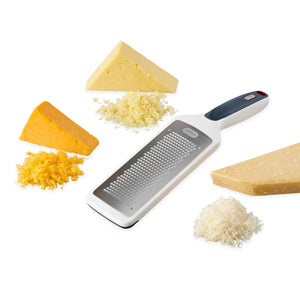 The Zyliss Smooth Glide Fine Grater, featuring a black handle, rests on a white surface amid wedges and shreds of yellow cheddar, white cheddar, and parmesan—ideal for grating cheese or zesting lemons. 