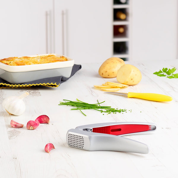 The Zyliss Susi 3 Garlic Press, in silver and red with an ergonomic handle, sits on a white kitchen counter among chopped chives, garlic cloves, potatoes, parsley, a yellow knife, and a baked dish. Its also dishwasher safe for easy cleanup. 