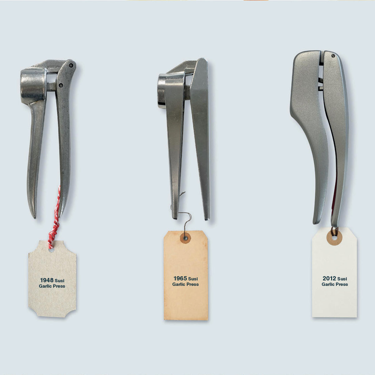 Three metal garlic presses are displayed vertically in a row, each with a tag: 1948 Swai Garlic Press, 1965 Swai Garlic Press, and 2012 Swai Garlic Press, showing their evolving design over time.