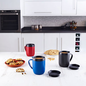 Three Zyliss Hot Mug Cafetière Mugs in blue, red, and black with double-wall insulation sit on a white kitchen table with cookies and bread. Two black lids are nearby. Modern white cabinets and appliances complete this coffee lovers scene. #grey
