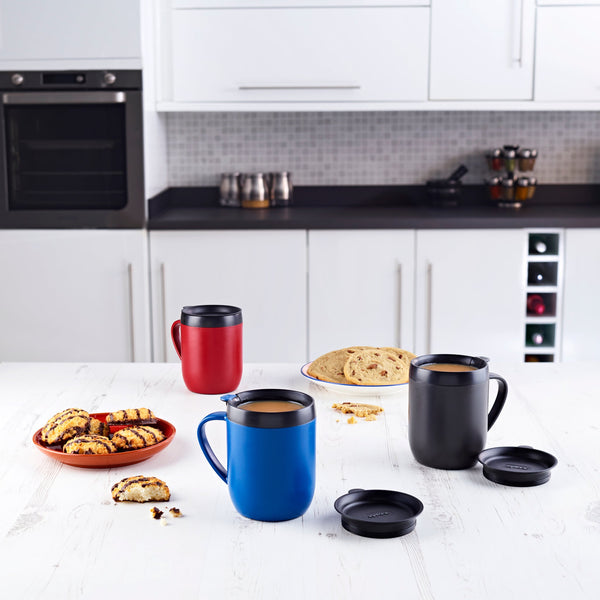 Three Zyliss Hot Mug Cafetière Mugs in red, blue, and black sit on a white kitchen table with plates of cookies and bread—ideal for coffee lovers. #blue