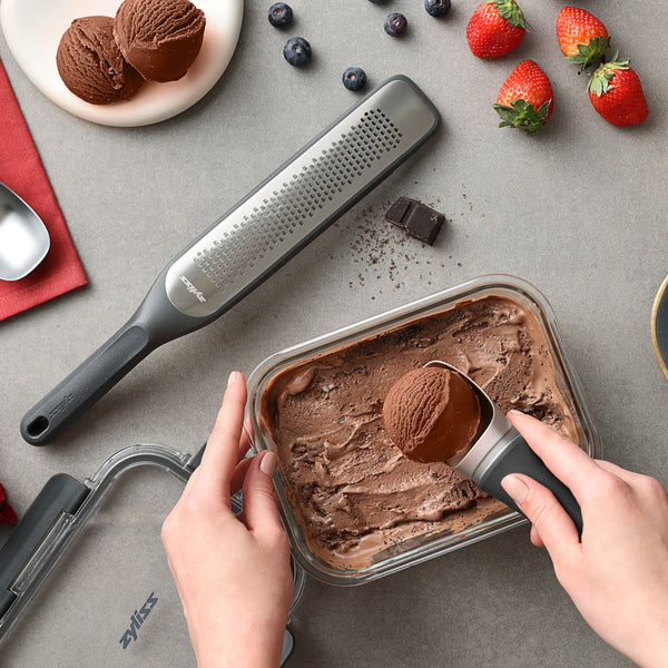 A person scoops chocolate ice cream from a container surrounded by a grater, chocolate pieces, blueberries, and strawberries—ideal for meal prep or storing leftovers with Zyliss Glass Container for secure, fresh storage. #0.37L/0.39qt