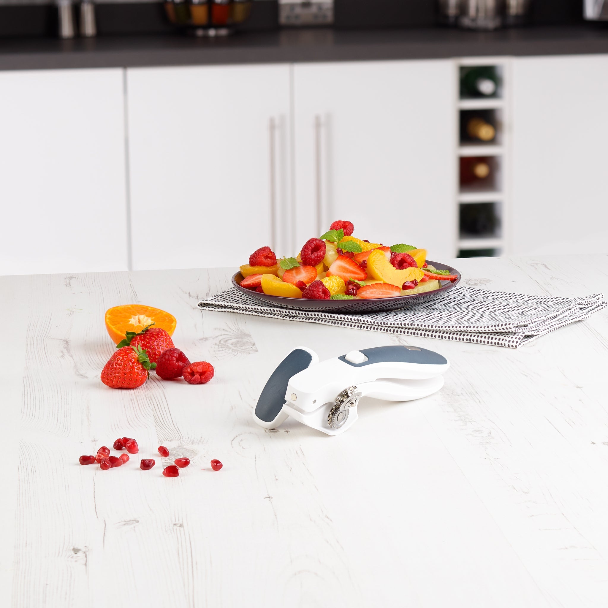 A plate of assorted fresh fruit sits on a folded napkin on a white kitchen counter, with the Zyliss Lock & Lift Can Opener, strawberries, an orange slice, and scattered pomegranate seeds in the foreground.
