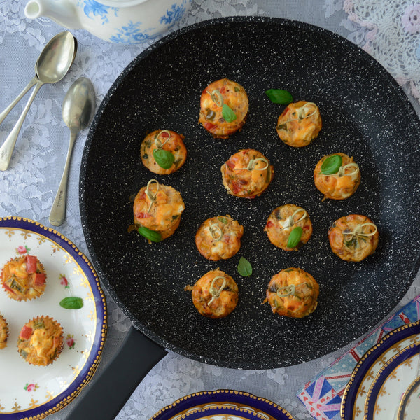 The Zyliss Ultimate Non-Stick Frying Pan in black holds savory, herb-topped muffins. It’s displayed on a lace tablecloth with vintage plates, a teapot, and spoons surrounding it. #24cm/9½"