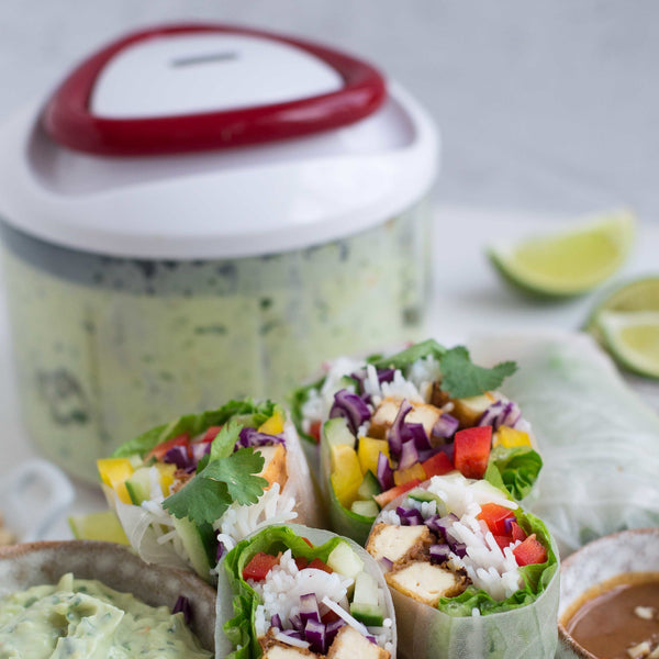 Fresh spring rolls with rice, tofu, vegetables, and cilantro are plated. The veggies are diced effortlessly using the Zyliss Easy Pull Food Processor. A green dip and lime wedges provide a fresh touch to this appetizing arrangement. 