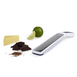 The Zyliss Smooth Glide Rasp Grater with a sharp blade rests on a white surface beside a cheese wedge, chocolate pieces with shavings, and a whole lime plus a lime wedge. 