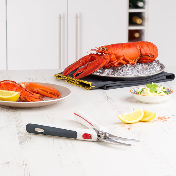 A whole cooked lobster sits on ice next to a plate of lobster meat, lemon wedges, butter, and Zyliss Shellfish Shears with stainless steel blades on a white kitchen counter. 