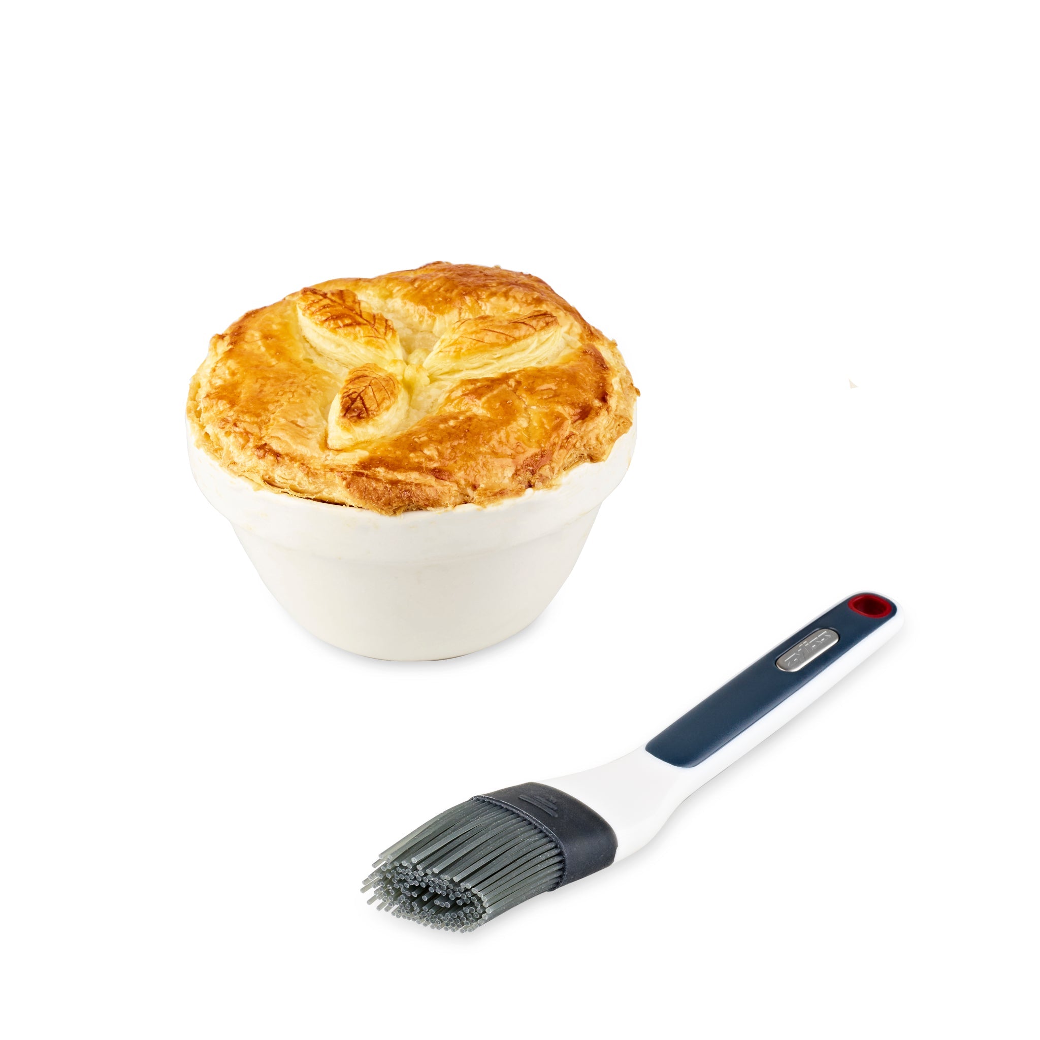 A golden-brown pot pie in a white bowl sits next to a Zyliss Silicone Pastry Brush with a black and gray handle, on a white background.