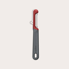 The Zyliss Soft Skin Peeler features a dark gray handle with a small end hole, a red top, and a horizontal stainless steel blade. This eco-friendly peeler is displayed on a plain light background. 