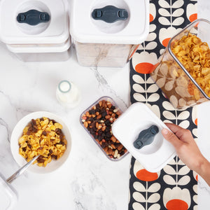 A hand opens a Zyliss Twist & Seal 5 Piece Container filled with mixed nuts and dried fruits on a white marble counter. Nearby are more Twist & Seal containers, a bowl of cereal with milk, a spoon, milk bottle, and patterned cloth. 
