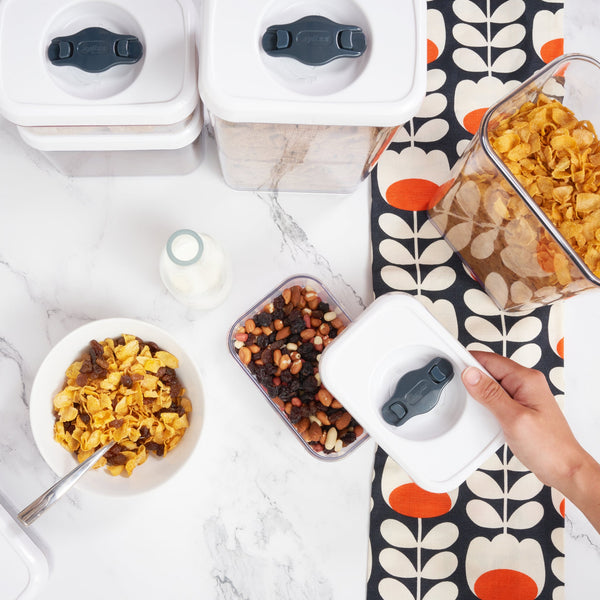 A hand opens a Zyliss Twist & Seal 5 Piece Container filled with mixed nuts and dried fruits on a white marble counter. Nearby are more Twist & Seal containers, a bowl of cereal with milk, a spoon, milk bottle, and patterned cloth. 