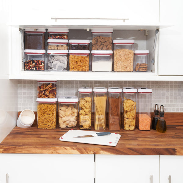 A kitchen countertop features the Zyliss Twist & Seal 5 Piece Container Set, efficiently storing dry foods like pasta, cereal, and nuts. Clear airtight containers with twist-lock lids, a cutting board, knife, olive oil bottles, and white canisters complete the scene. 