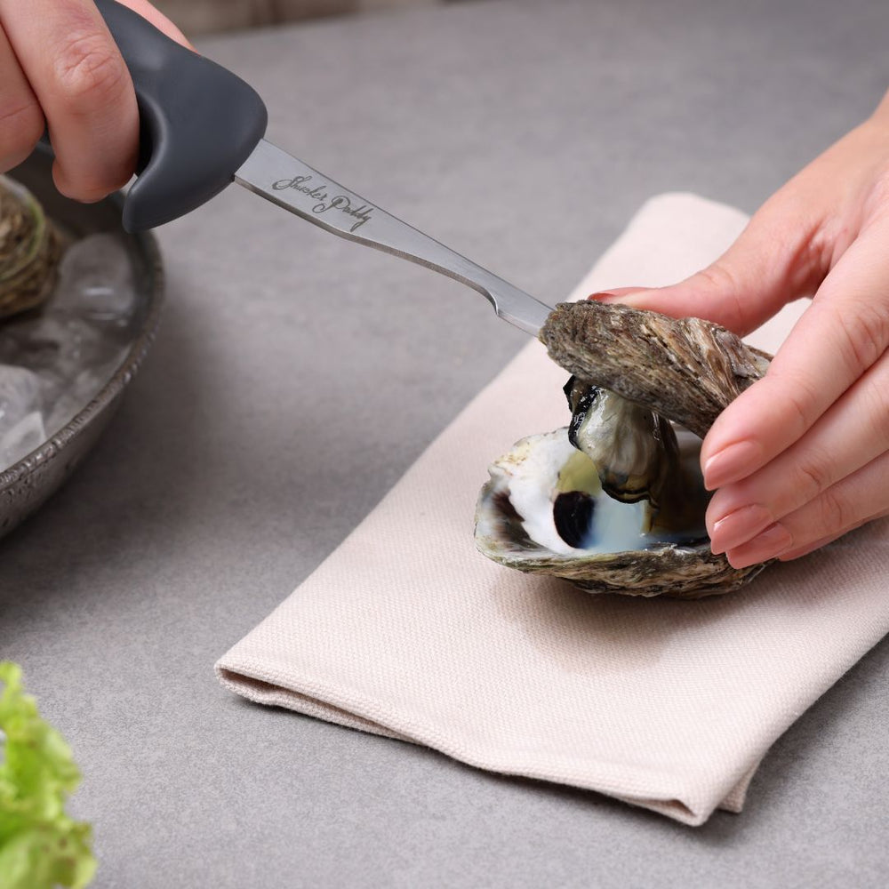 The Zyliss Kitchen Oyster Knife Shucker Paddy, featuring a hooked end, ergonomic black handle, and Shucker Paddy signature on the blade, is displayed against a plain white background.