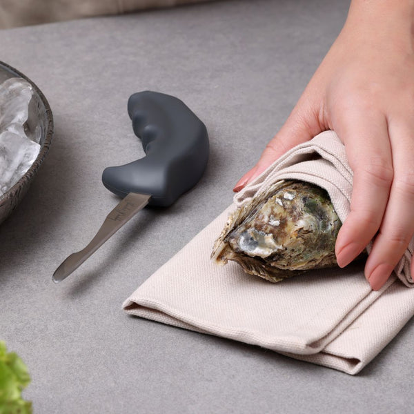 A hand prepares to shuck an oyster wrapped in a cloth using the Zyliss Kitchen Oyster Knife Shucker Paddy on a gray countertop beside a bowl of ice.