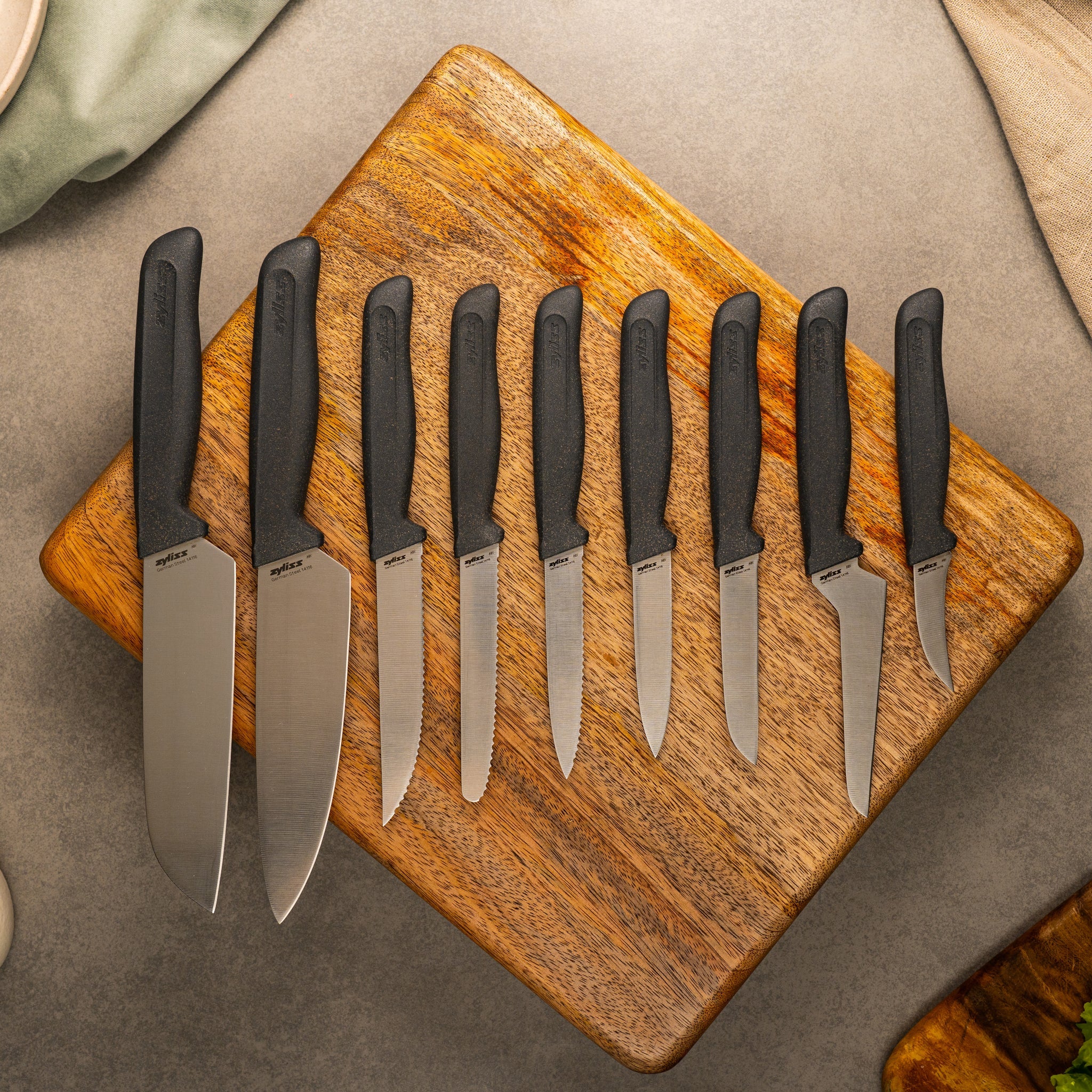 Eight kitchen knives with black handles are neatly arranged in a row on a wooden cutting board, displayed from largest to smallest blade, on a stone countertop with fabric napkins in the corners.